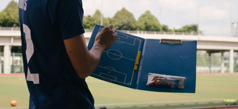 Ein Trainer hält ein Taktikboard mit einem Fußballfeld und Notizen in der Hand.
